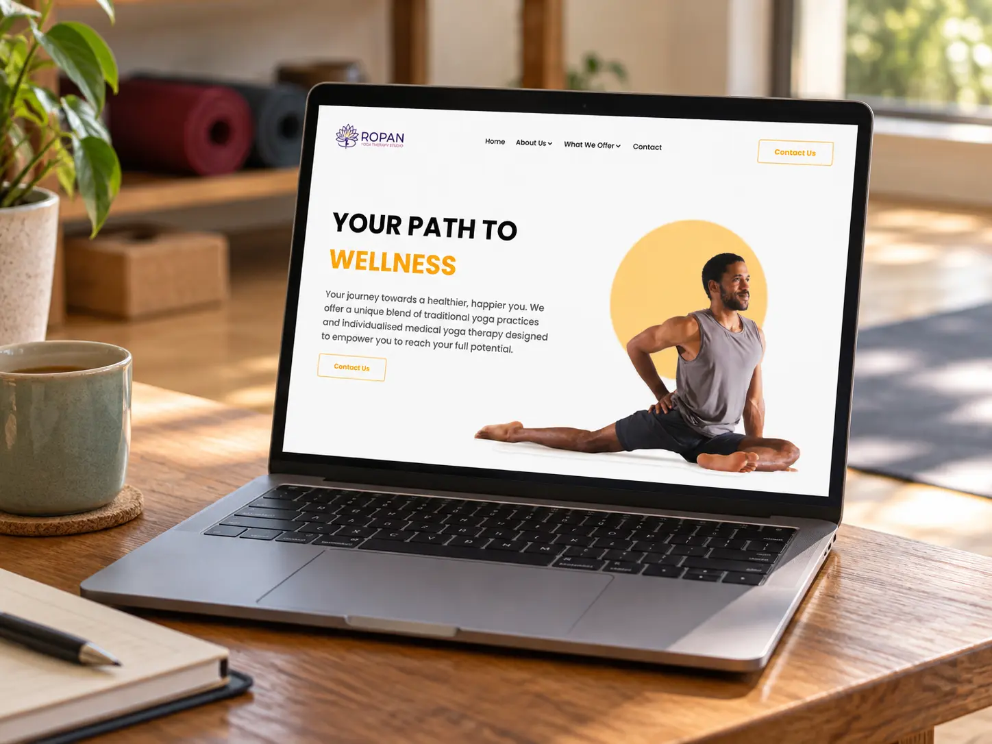 A laptop on a low wooden table in a sunlit studio, showing Ropan's landing page: 'Your path to wellness' in black and amber.
