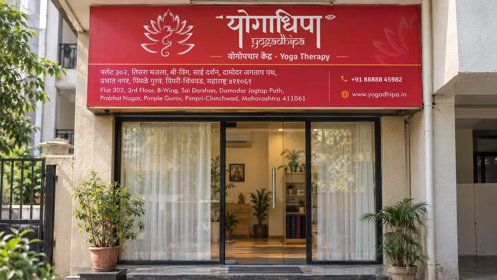 The crimson bilingual Yogadhipa fascia above the glass door of a small studio: the white lotus and योगाधिपा, 'योगोपचार केंद्र · Yoga Therapy', and a lower strip with the address in Marathi and English, the phone number and the website; a tulsi plant by the step.